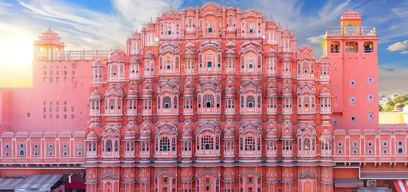 Daytime view of the ornate pink sandstone facade and windows of Hawa Mahal in Jaipur under a blue and cloudy sky