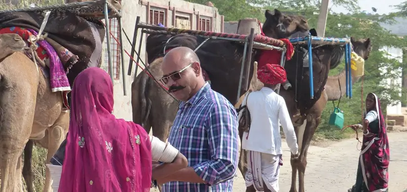 Villagers interacting near pack camels laden with goods in a traditional Indian desert village.